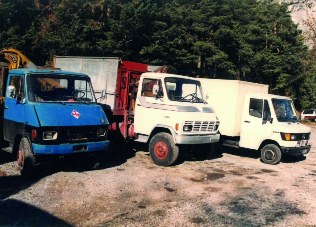 Drei alte Lieferwagen von Unternehmen, geparkt auf Schotter, mit Bäumen im Hintergrund.