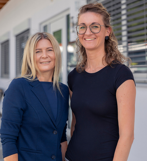 Zwei Frauen des Tschiderer-Teams lächeln vor einem Gebäude mit Fenstern und Jalousien.