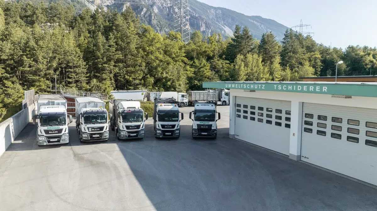 Fünf Lastwagen aus dem Tschiderer Fuhrpark parken vor einer Garage mit der Aufschrift Umweltschutz in der Nähe eines Waldes.